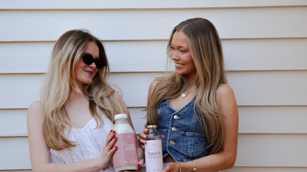 Two women stand in front of a white wall, smiling and holding bottles labeled “Milk” and “Ranch Water.”.