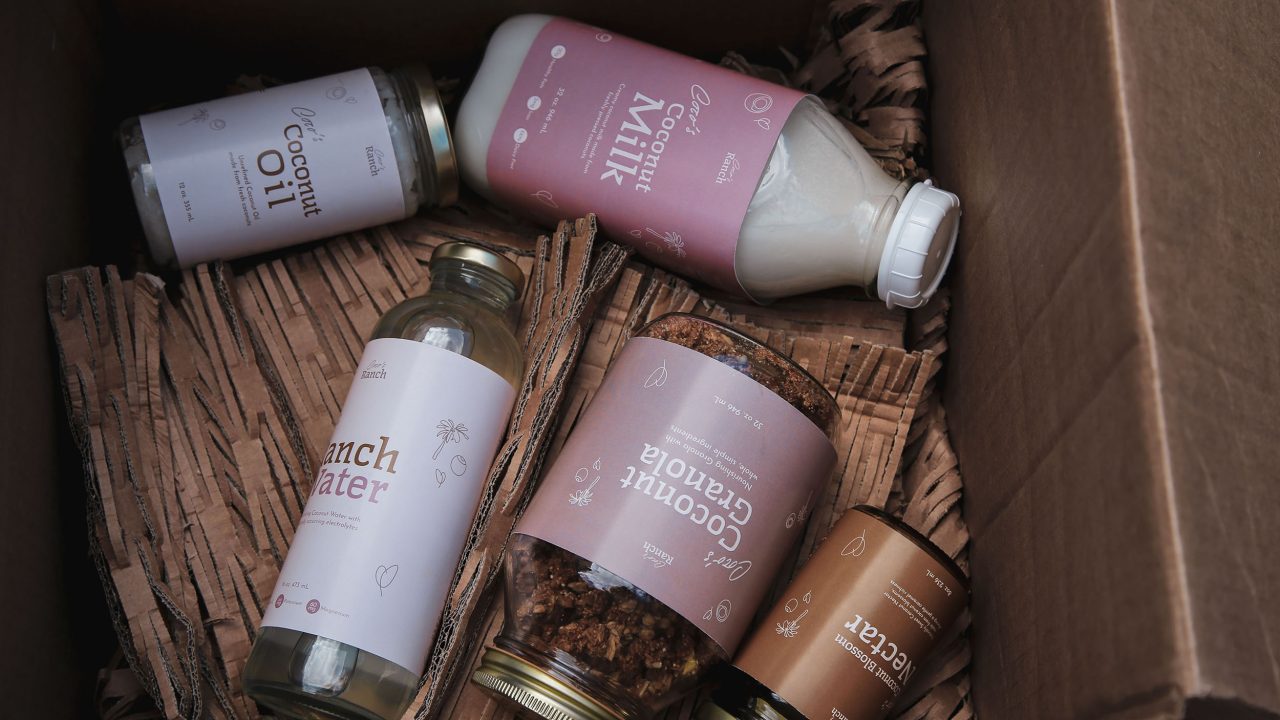 A cardboard box containing bottles and jars labeled coconut milk, coconut cordial, coconut granola, beach water, and nectar, arranged on brown packaging material.