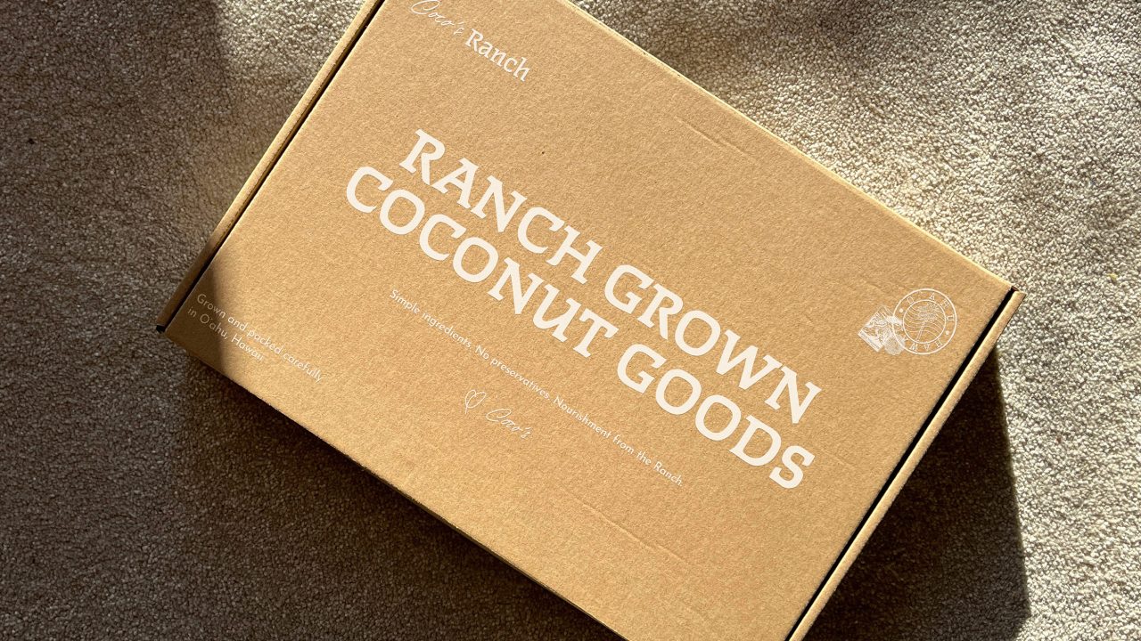 A brown cardboard box on carpet, labeled Ranch Grown Coconut Goods in large white letters with smaller text and simple graphics on the lid.