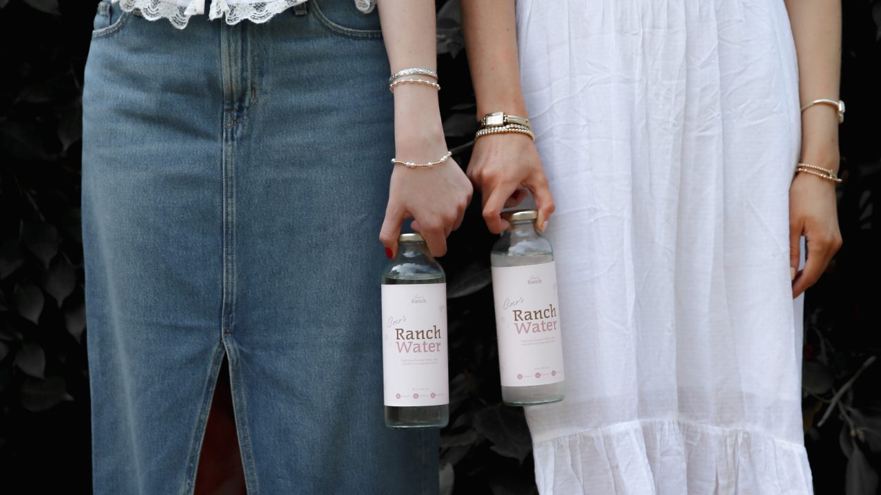 Two women stand side by side outdoors, each holding a can of Ranch Water. They wear denim and white outfits with cowboy boots, and their faces are not visible.