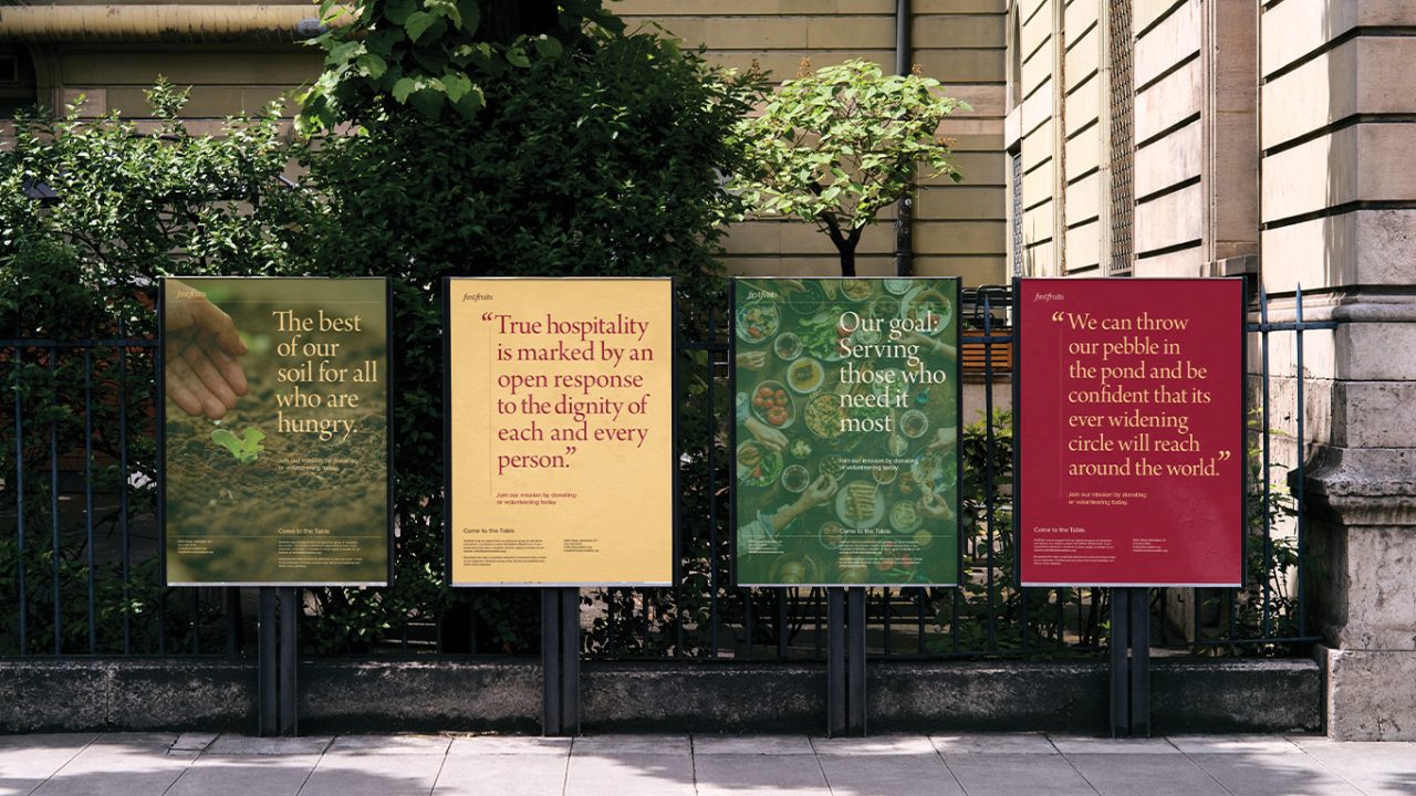 Four colorful posters with inspirational quotes about hospitality and service are displayed on stands along a sidewalk, in front of a metal fence and greenery.