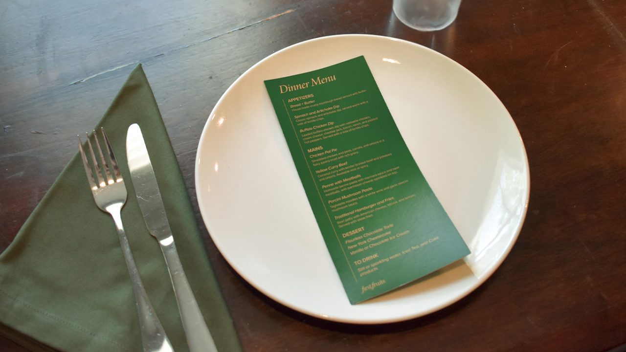 A green dinner menu is placed on a white plate with a fork, knife, and green napkin on the left, set on a dark wooden table.