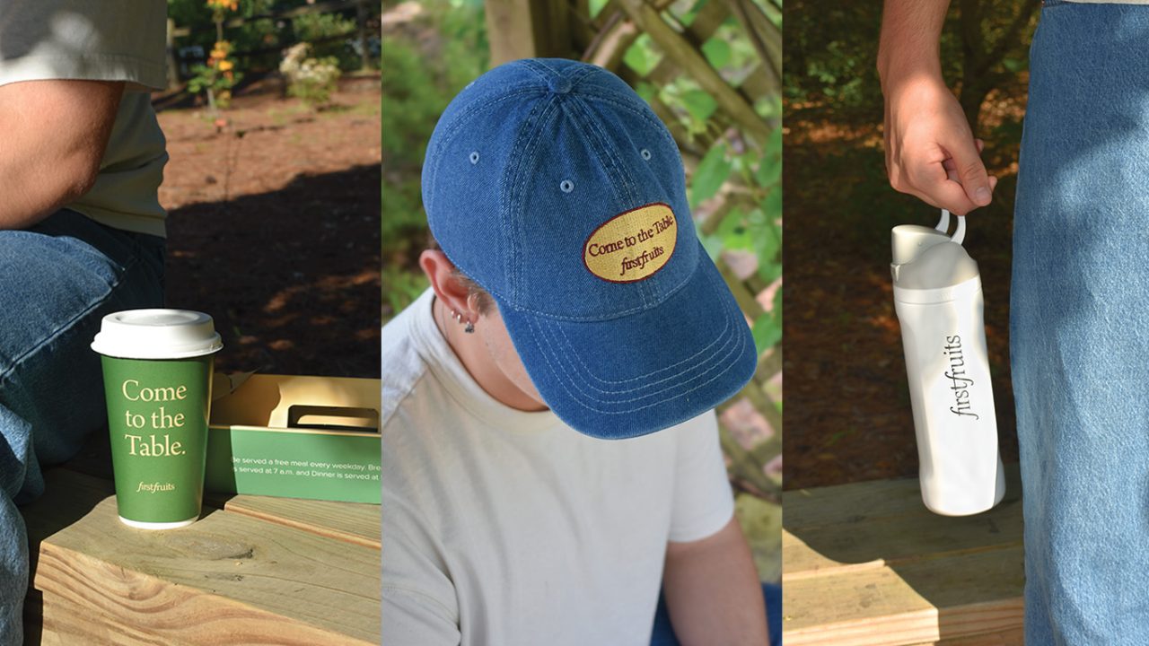 A person with a coffee cup, a blue Come to the Table cap, and a white firstfruits water bottle, all featuring branded logos, sits outdoors on a wooden bench.