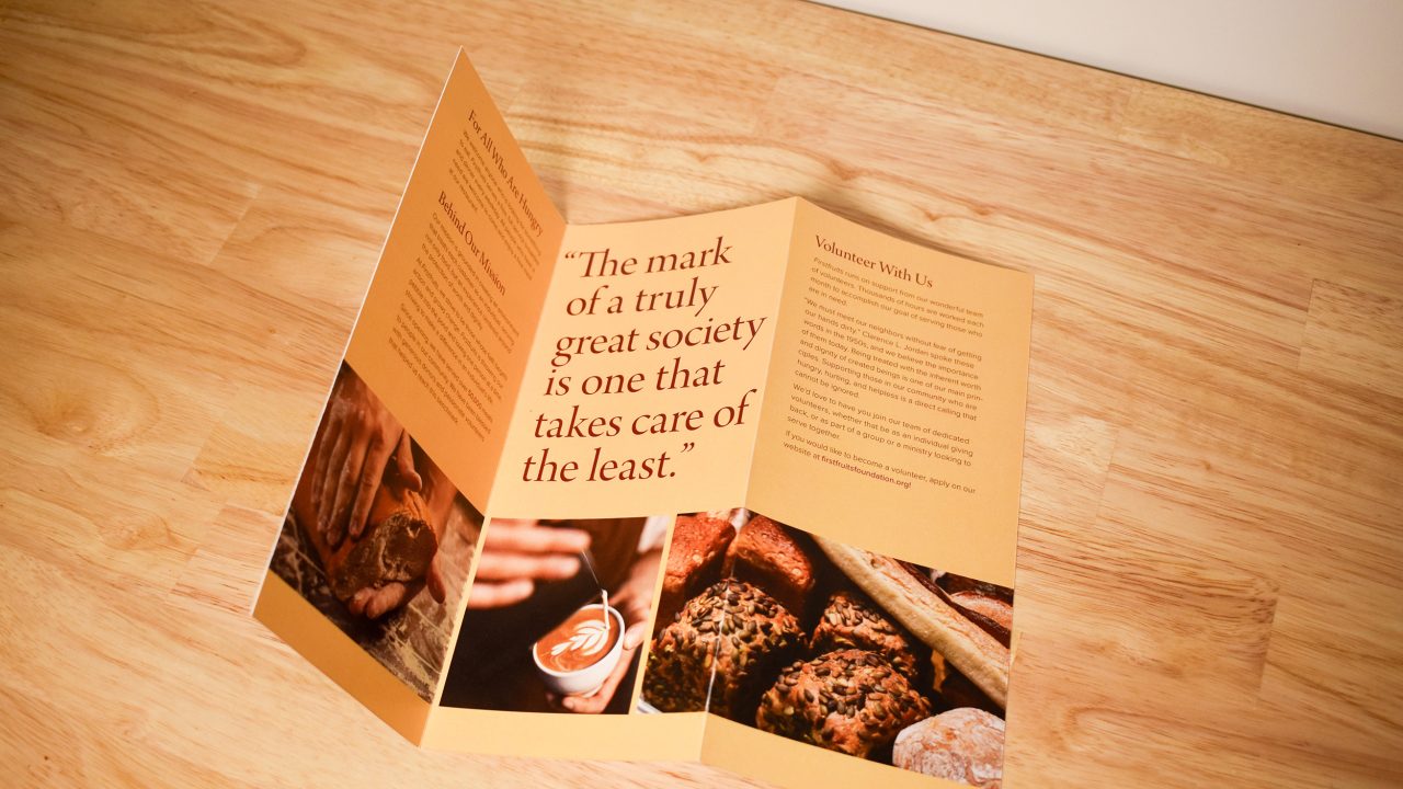 A trifold brochure with text and images about volunteering is displayed on a wooden surface, highlighting a large quote and photos of hands, bread, and people.