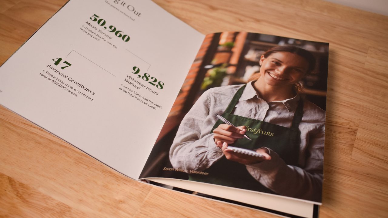 Open booklet on a wooden table showing a woman in an apron holding a notepad on the right page, and statistics about volunteers and contributions on the left page.