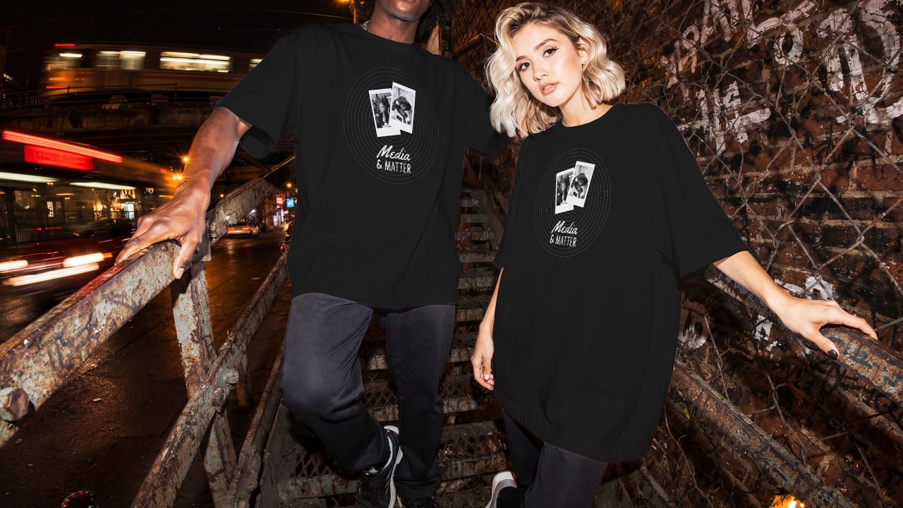 Two people wearing oversized black graphic t-shirts stand on an outdoor metal staircase at night, with graffiti on the walls and blurred city lights in the background.