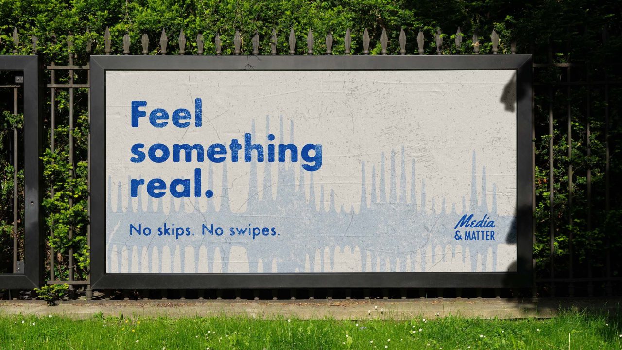 A billboard on a black metal fence displays the text: Feel something real. No skips. No swipes. Media & Matter, with a faint abstract background. Trees and grass are visible nearby.