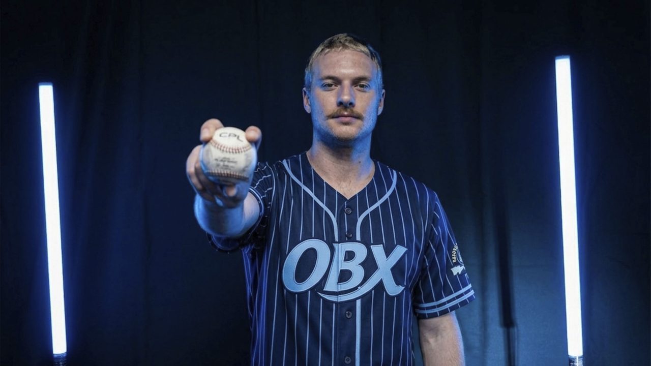 A baseball player in a striped OBX jersey holds a baseball toward the camera, standing between two vertical blue lights against a dark background.