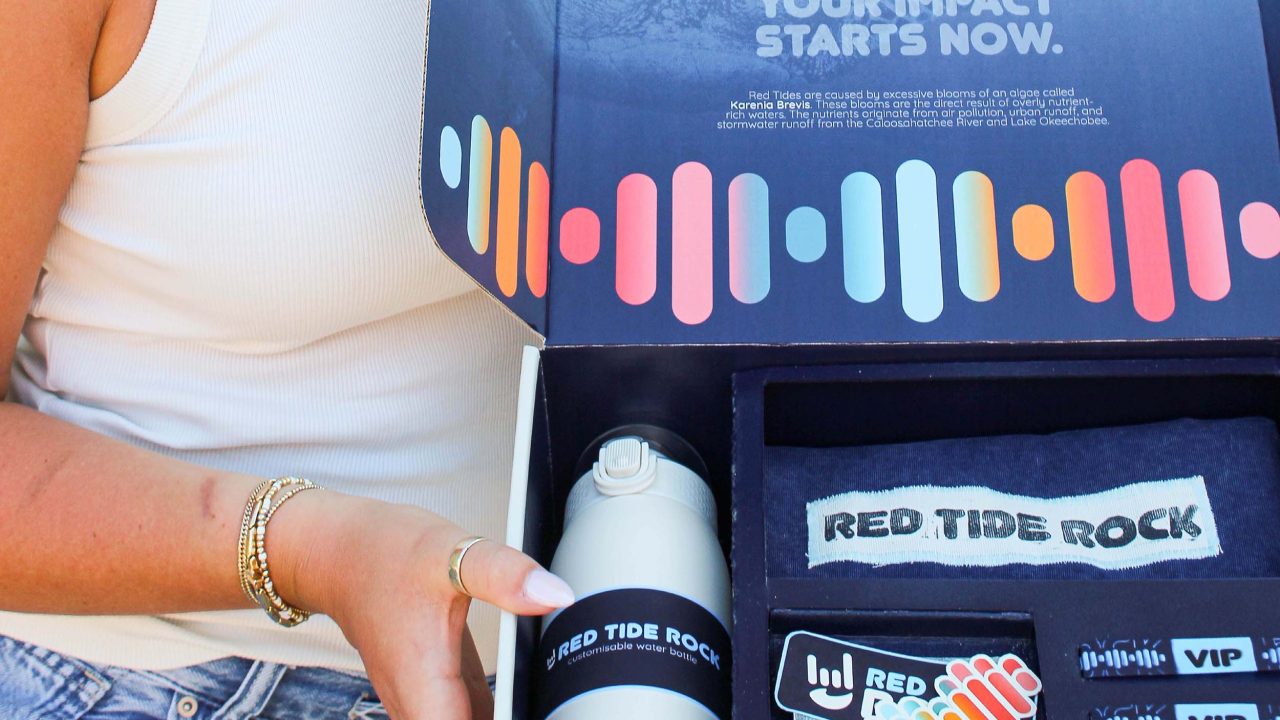 A person holds an open box containing a water bottle, T-shirt, stickers, and a card. The inside lid reads, YOUR IMPACT STARTS NOW with colorful graphics.