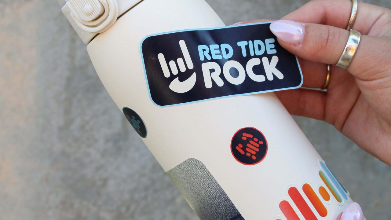 A hand places a RED TIDE ROCK sticker on a white water bottle already decorated with colorful and black circular stickers.