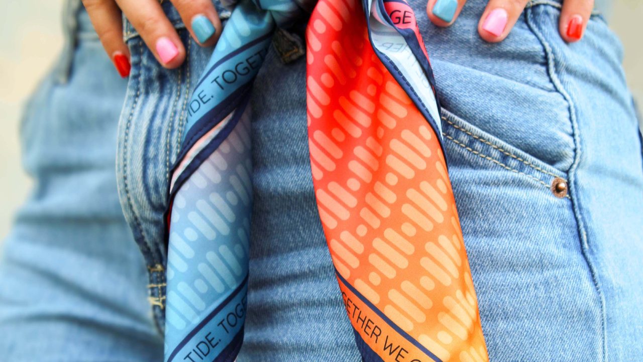 Close-up of a person wearing blue jeans with a colorful scarf tied at the waist. The person has red, blue, and pink nail polish on their fingernails.