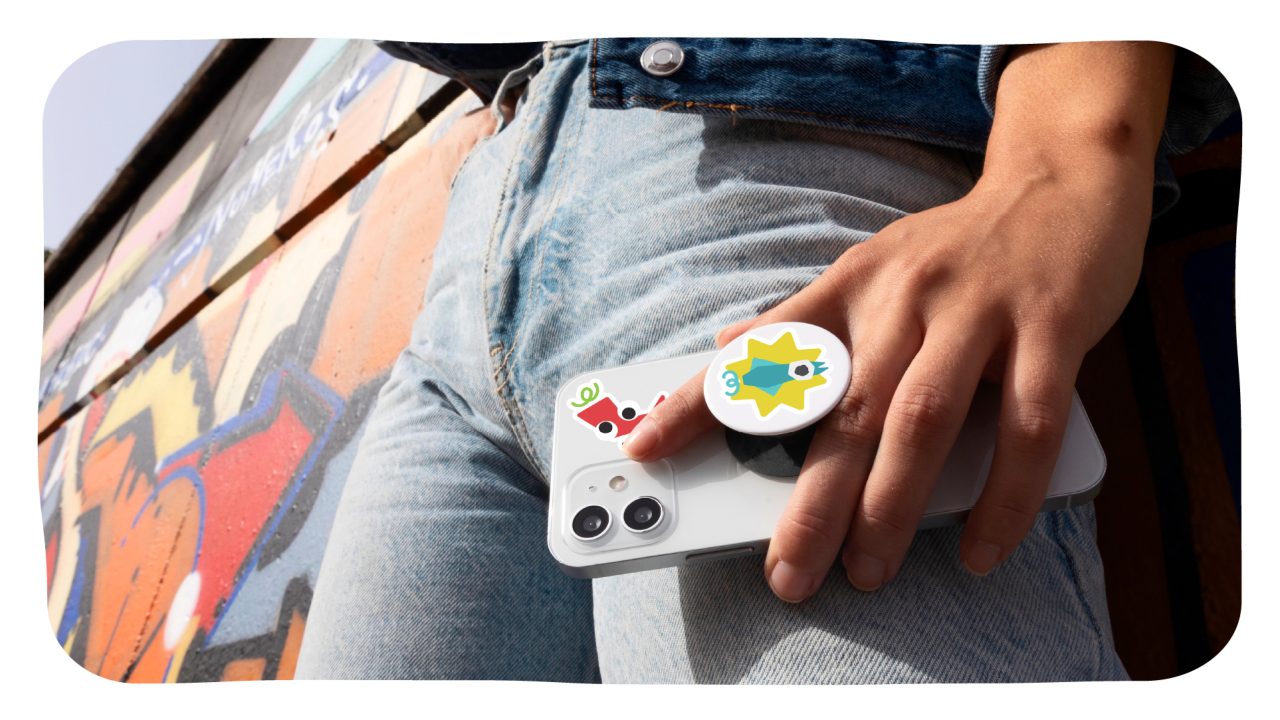 Person in blue jeans holds a white smartphone with a pop socket and stickers, standing in front of a colorful graffiti wall.