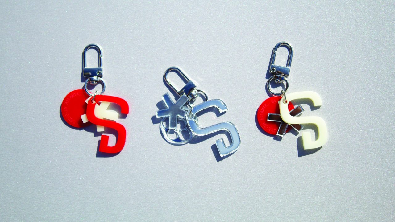 Three keychains with the letter S in red, clear, and white, each attached to a metal clasp and a round red tag, are displayed on a light gray surface.