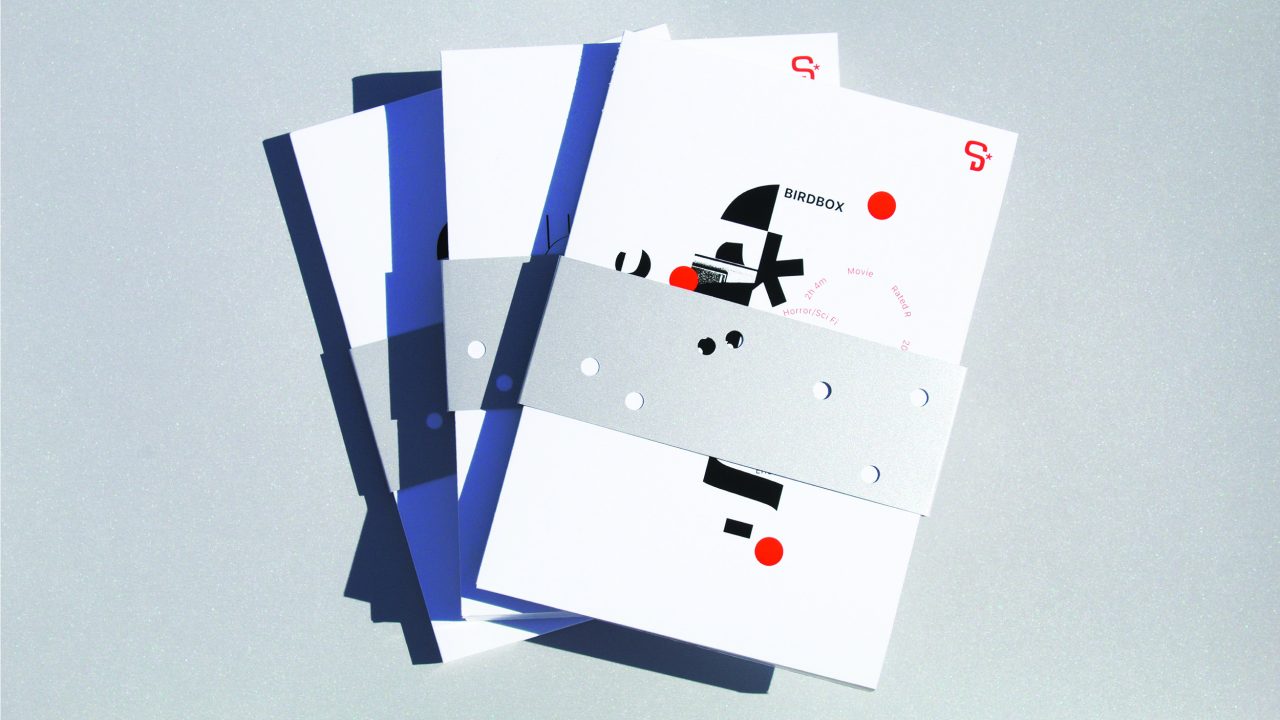 A stack of white booklets with minimalist abstract designs and red dots, held together by a metallic silver band with circular cutouts, on a light gray background.