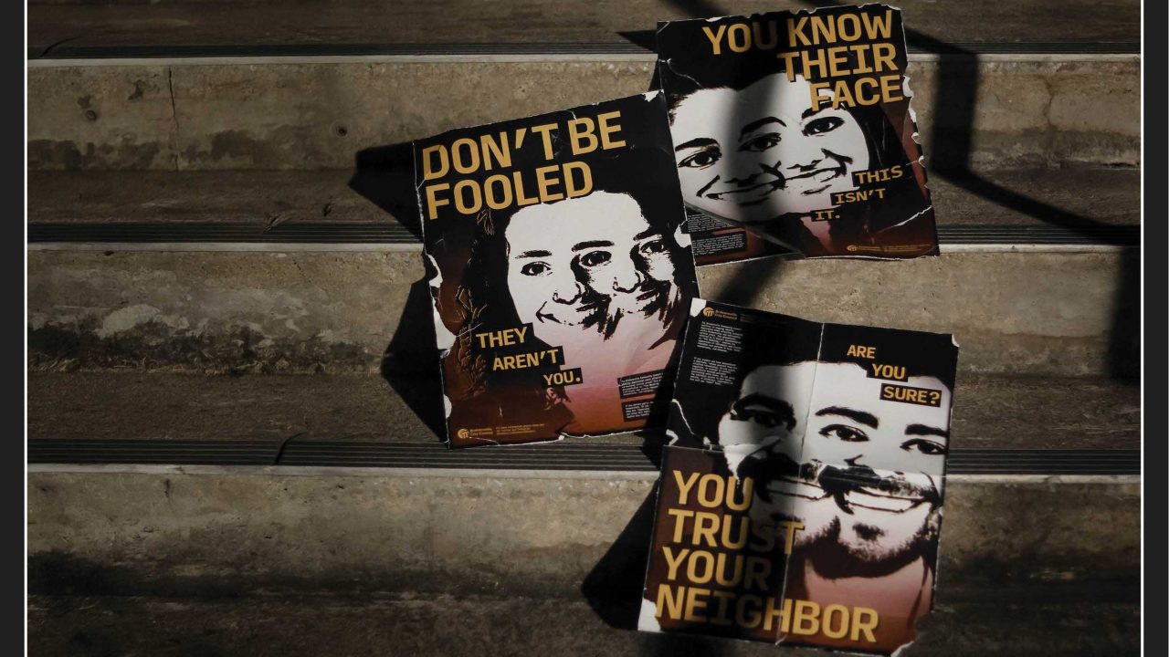 Three torn posters with smiling faces and bold warning messages, including DONT BE FOOLED and YOU TRUST YOUR NEIGHBOR, are scattered on concrete steps.