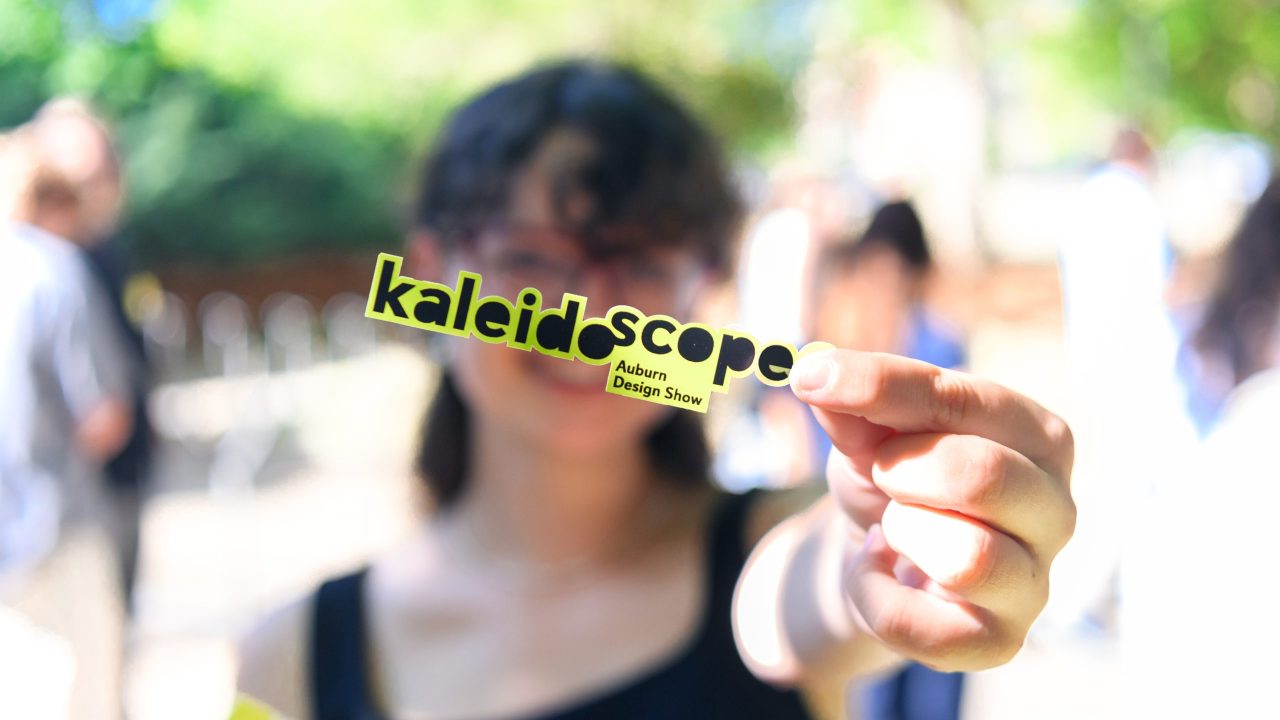 A person holds a yellow sticker reading kaleidoscope Auburn Design Show outdoors, with a blurred background of people and trees.