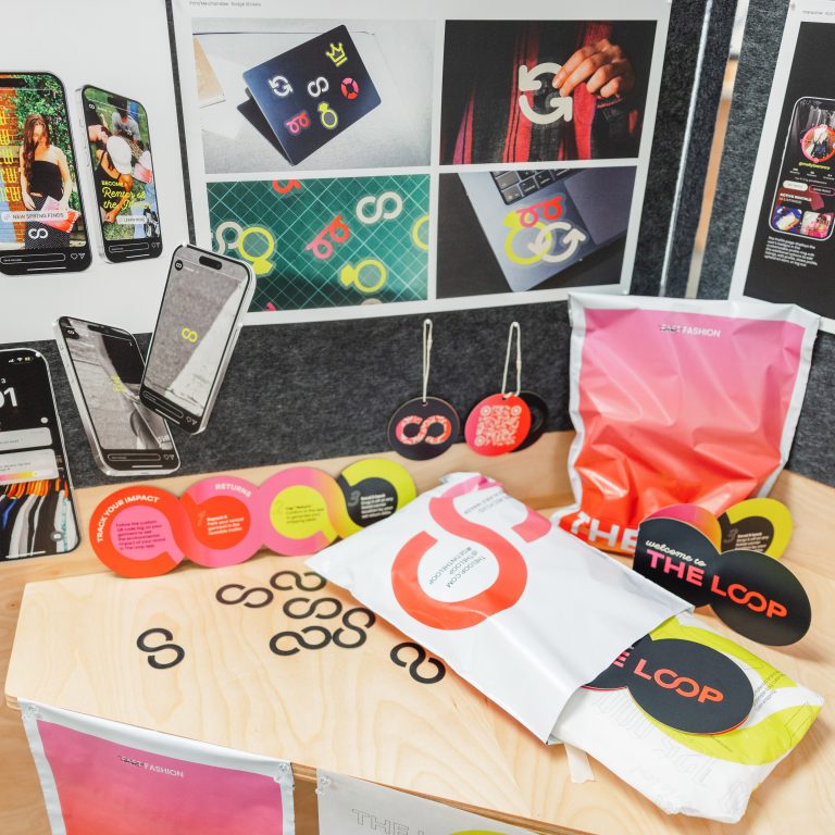 Assorted branded materials, including phone mockups, bags, buttons, and printed collateral, are displayed on a wooden table beneath marketing posters on a wall.