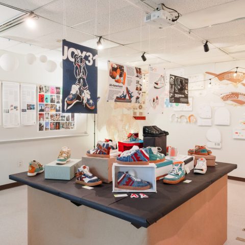 Display of colorful sneakers on a central table in a well-lit exhibition room, with posters, information panels, and hanging decorations on the walls and ceiling.