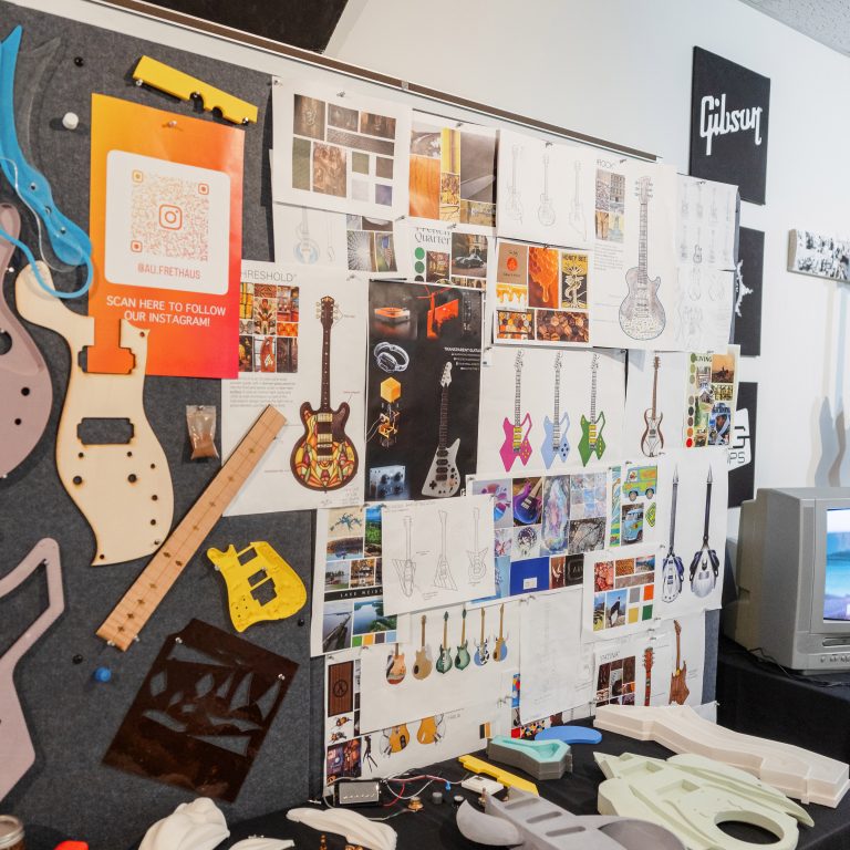 A workspace with guitar body parts, tools, and materials on the table; a wall behind displays sketches, photos, and designs of guitars and related reference images.
