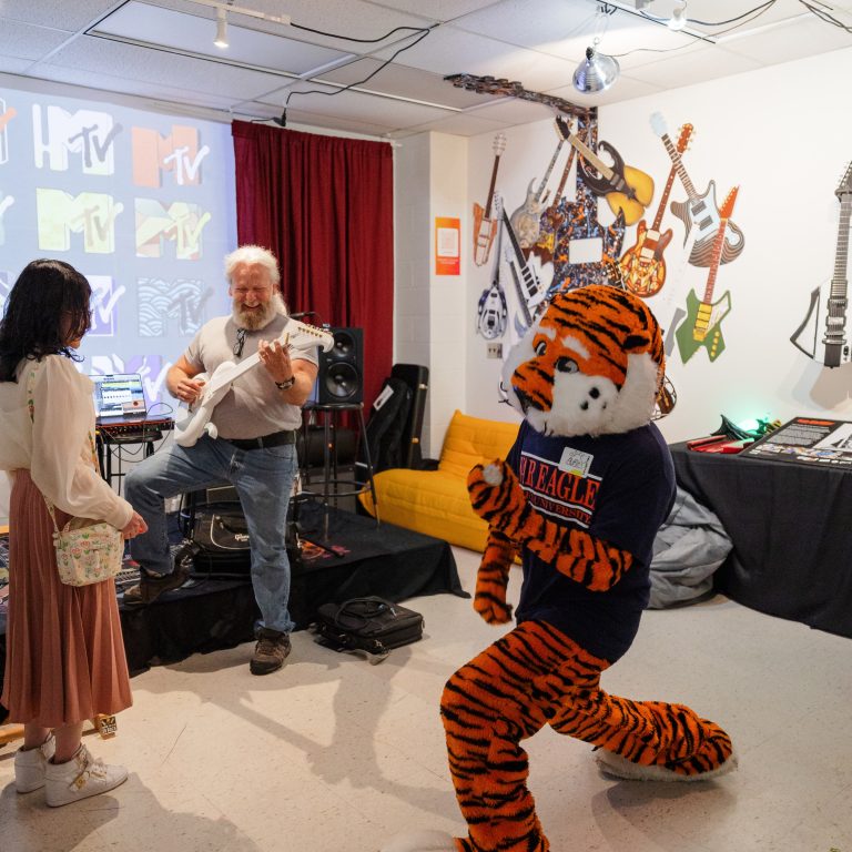 A person in a tiger mascot costume poses in the foreground while two people play guitar in a decorated room with MTV logos and guitars on the wall.