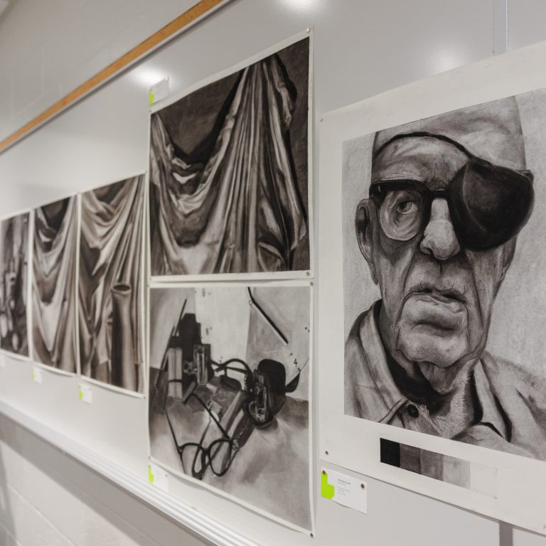 Charcoal drawings displayed on a whiteboard: draped fabric studies, a still life with headphones, and a portrait of an older man wearing an eye patch and glasses.