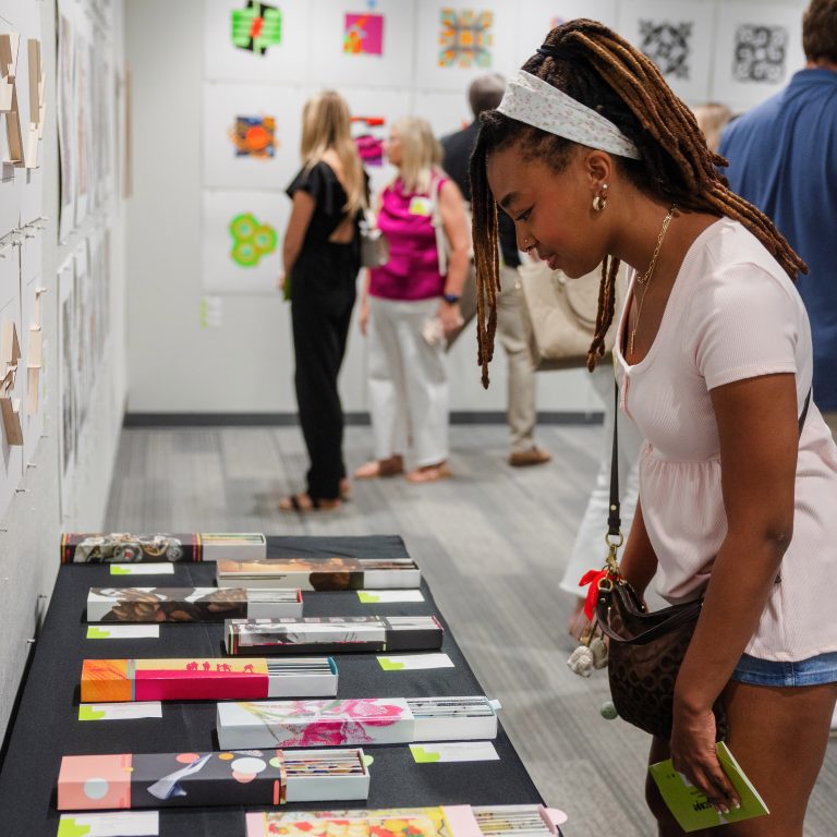 A woman examines colorful artwork displayed on a table at an art exhibit, with additional people viewing pieces on the walls in the background.