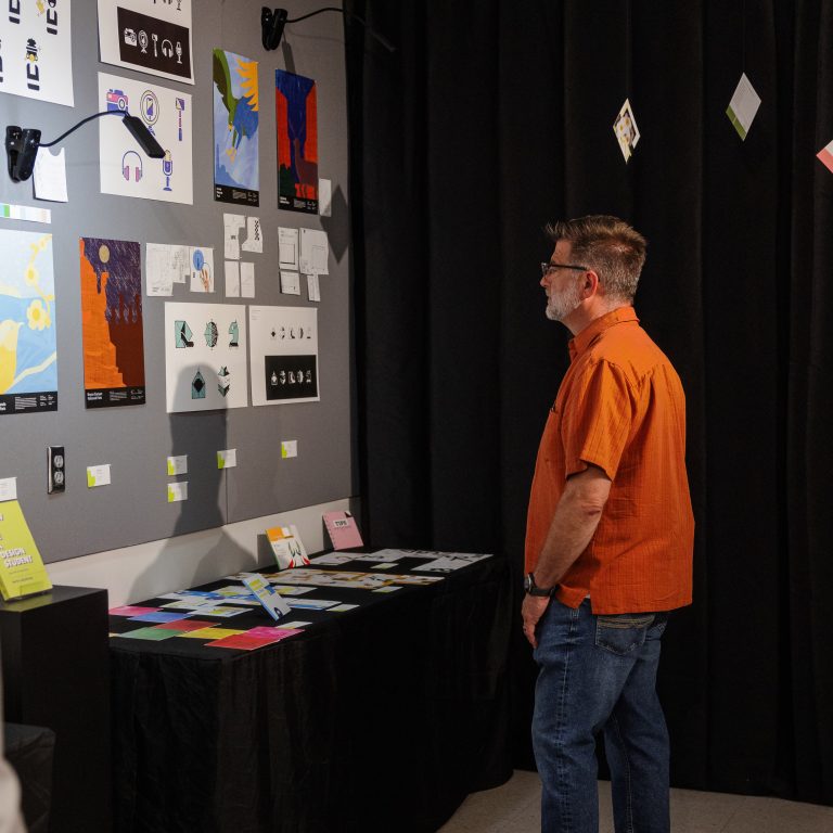 A man in an orange shirt stands in front of a wall displaying various artworks and designs in a gallery or exhibition space.