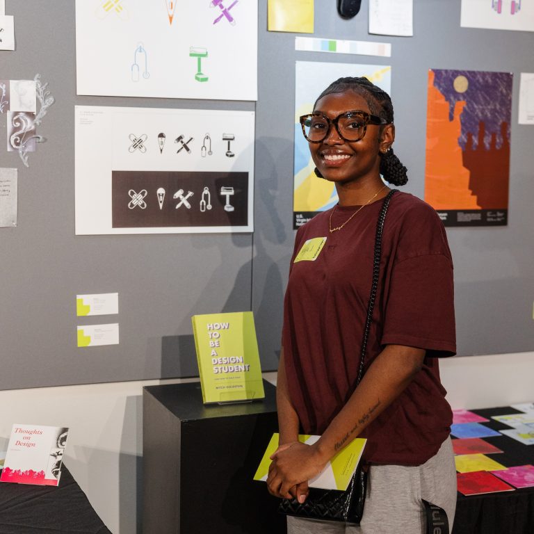 A person wearing glasses and a maroon shirt stands smiling in front of a display of graphic design artworks and posters on a wall.