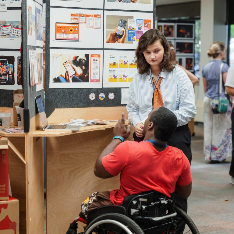 A woman stands by a display board with design materials, talking to a man in a wheelchair who gestures with his hand in a public indoor setting.