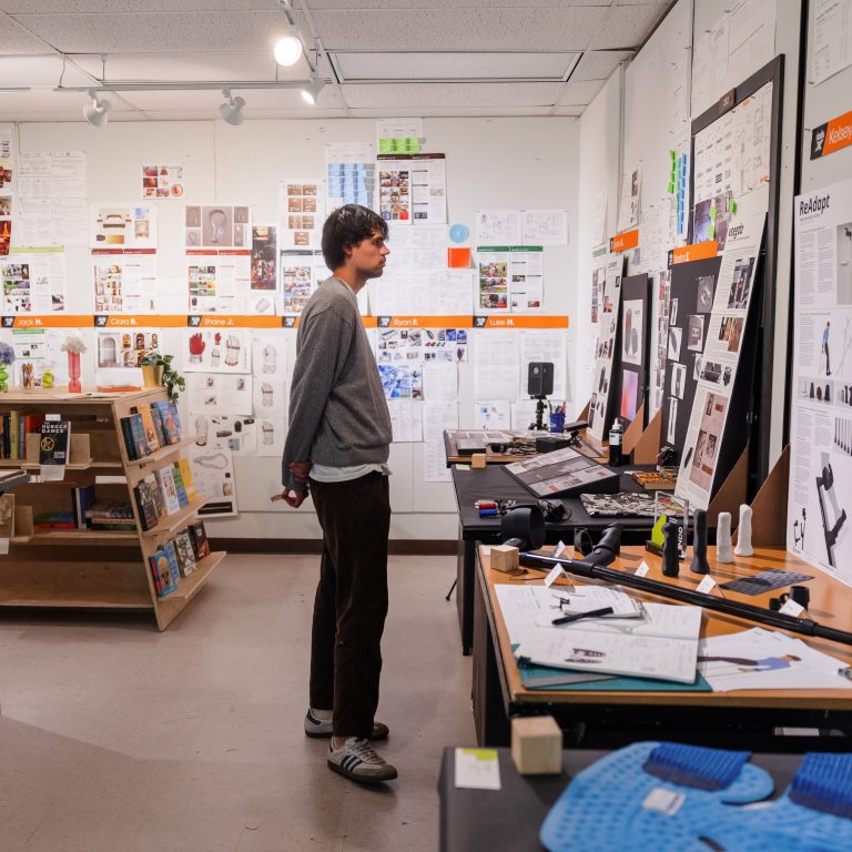 A person stands in a design studio, looking at walls covered with sketches, notes, and design plans. Several desks hold models, papers, and drawing tools.