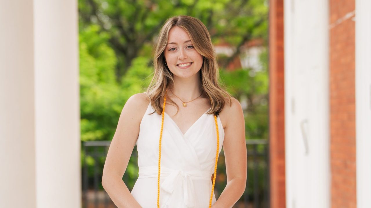 Young woman in a white dress stands on a marble walkway holding a graduation cap, wearing an honor cord, with green trees and columns in the background.