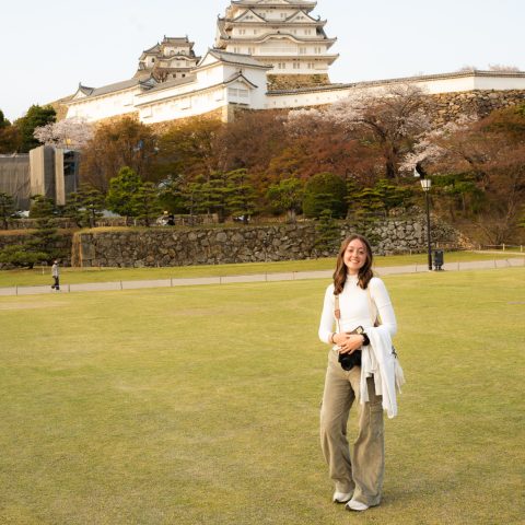 A person stands smiling on a grassy area with Himeji Castle in the background under a partly cloudy sky.