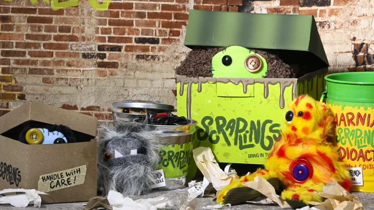 Several furry, colorful toy monsters with button eyes are displayed in and around green boxes and cans labeled Scrapkins against a brick wall background with scattered paper.