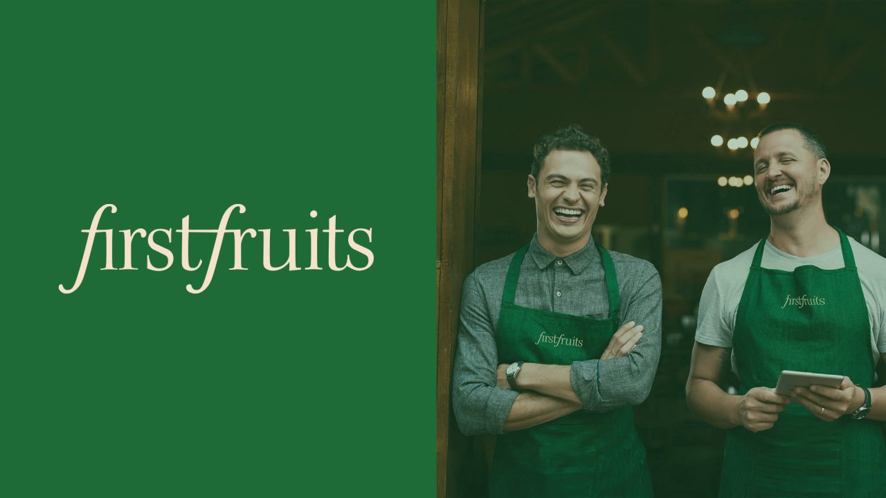 Two men in green aprons with firstfruits logo stand smiling indoors; the left side of the image displays the firstfruits logo on a solid green background.