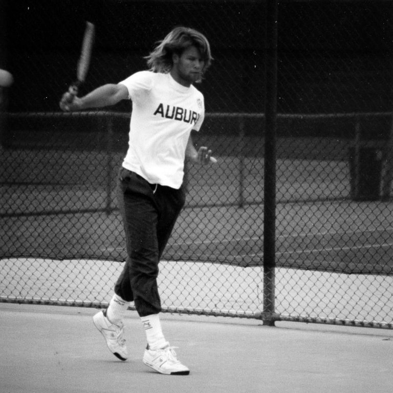 A person wearing an Auburn T-shirt plays tennis on an outdoor court, mid-swing, with a chain-link fence in the background.