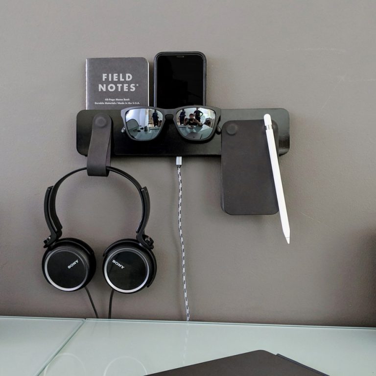 Wall-mounted organizer holding a phone, notebook, sunglasses, pen, and headphones above a glass desk with office supplies, a clock, and a closed laptop.