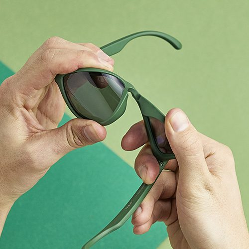 A person holds green sunglasses with both hands against a two-tone green background.