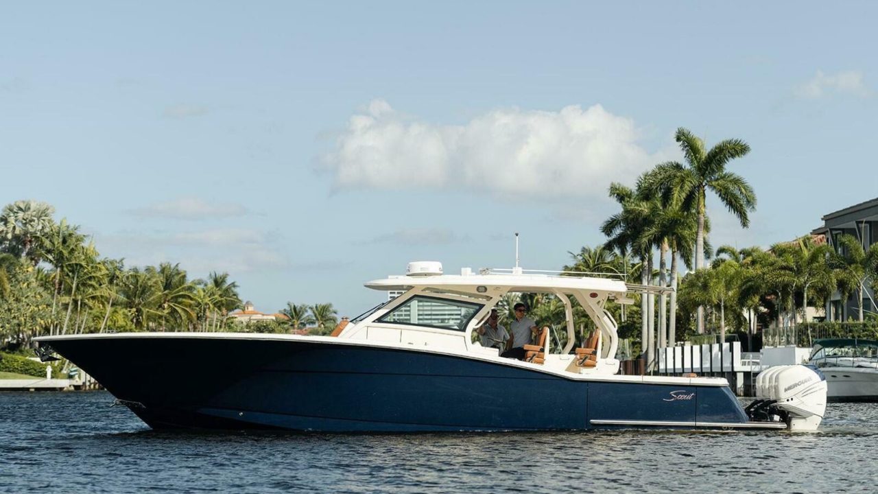 A dark blue and white motorboat with two outboard engines floats on a calm waterway, with palm trees and houses visible in the background.