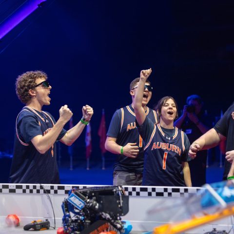 Five students wearing Auburn jerseys cheer and celebrate behind a robotics competition field, with a robot and game pieces visible in the foreground.