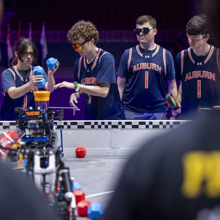 A group of students in Auburn jerseys operate a robot on a game field, interacting with blue and red blocks during a robotics competition.