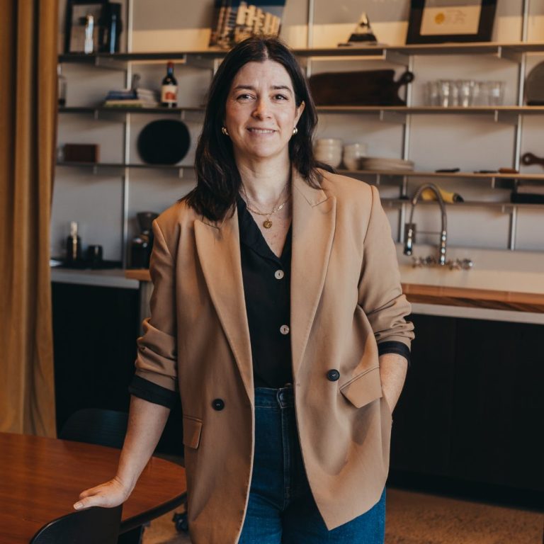 A woman stands next to a wooden table in a modern room with shelves displaying bottles and decor items, wearing a tan blazer, black top, blue jeans, and black shoes.