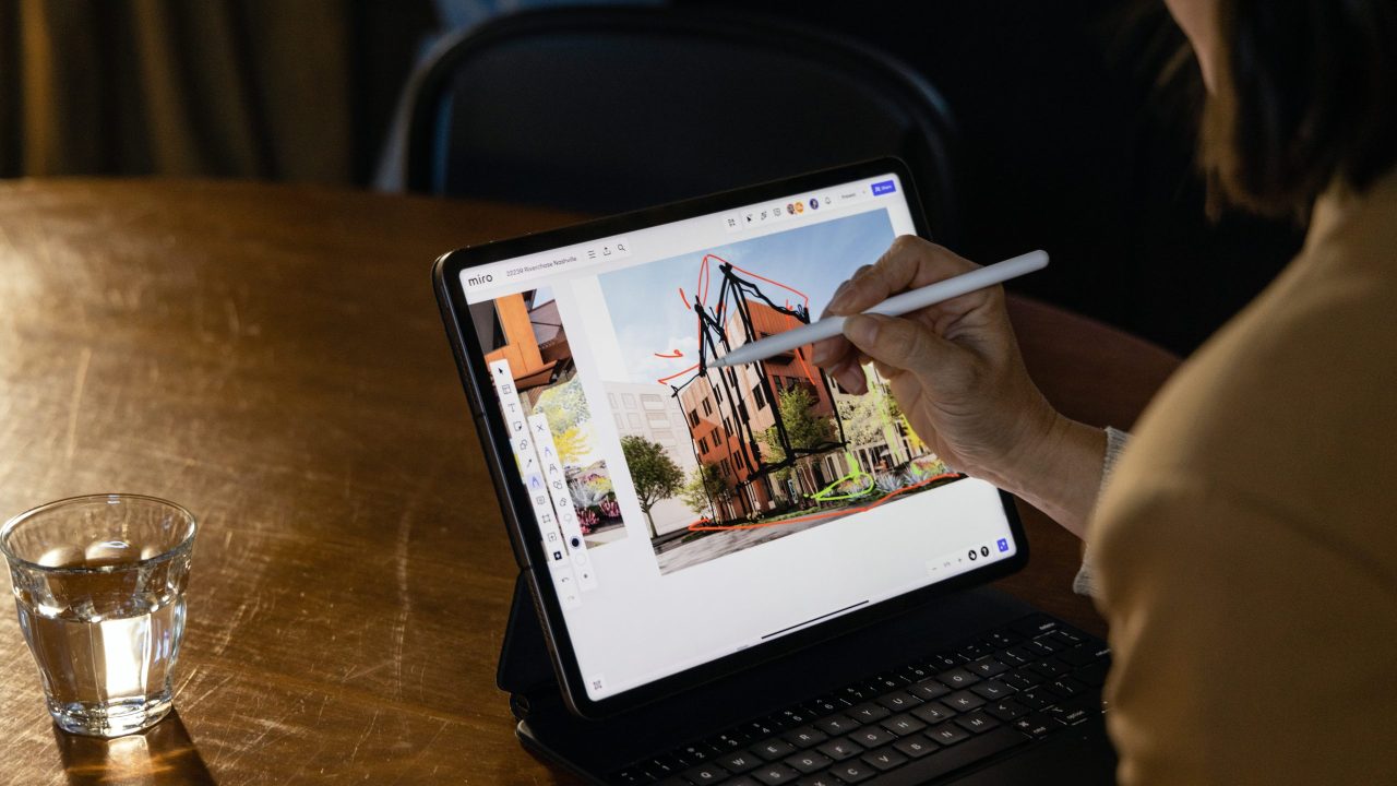 Person using a stylus to draw architectural designs on a tablet with a keyboard, seated at a wooden table with a glass of water nearby.