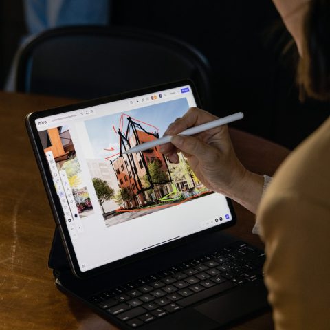 Person using a stylus to draw architectural designs on a tablet with a keyboard, seated at a wooden table with a glass of water nearby.