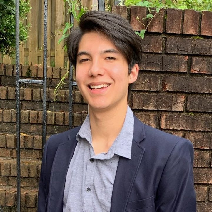 A young person stands outdoors in front of a brick wall and metal gate, wearing a navy blazer, grey shirt, light-colored pants, and black shoes, with hands in pockets.
