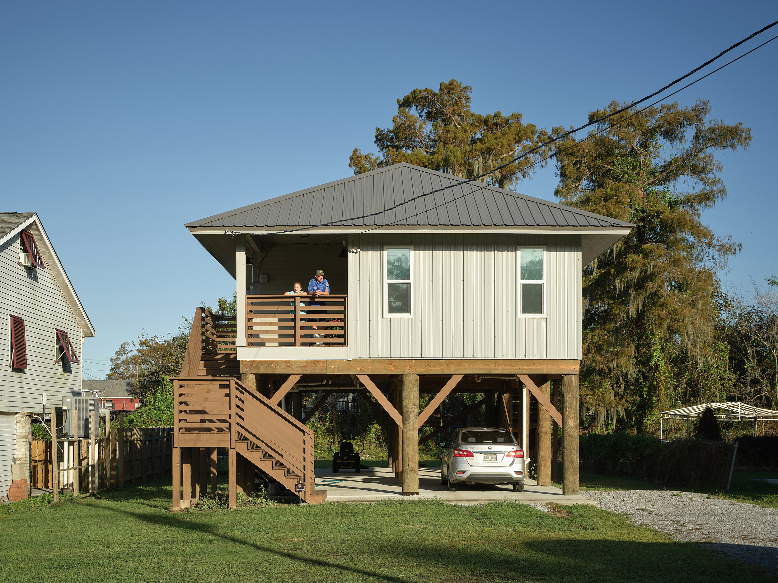 Elevated house on wooden stilts with a staircase, a porch with two people, and a car parked underneath on a sunny day.