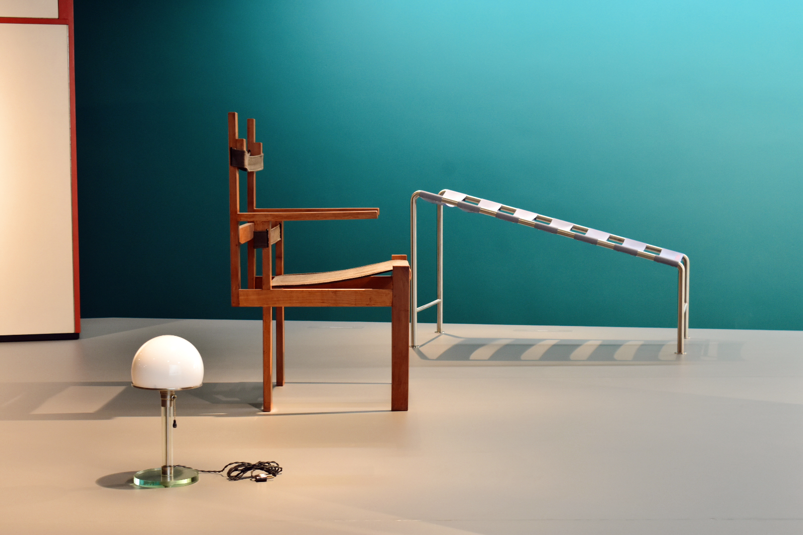 Wooden chair, metal slide-like structure, white dome lamp with cord, and teal background arranged in a minimalistic setting.