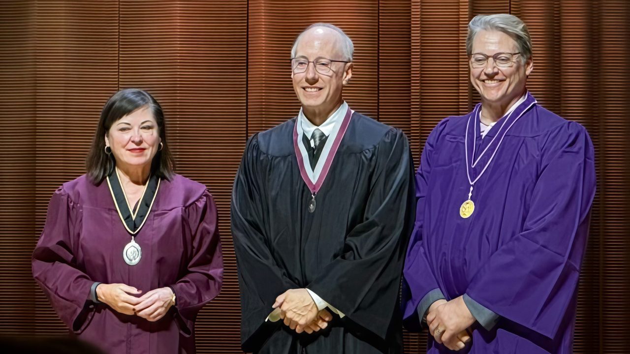 Three people in ceremonial robes and medals stand on stage in front of a brown curtain, posing for a photo.
