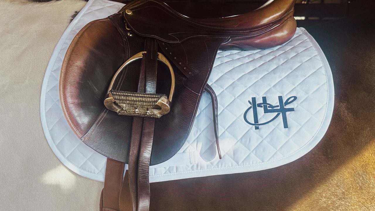 A brown English saddle on a white quilted saddle pad with a black logo, placed on a horse inside a stable.