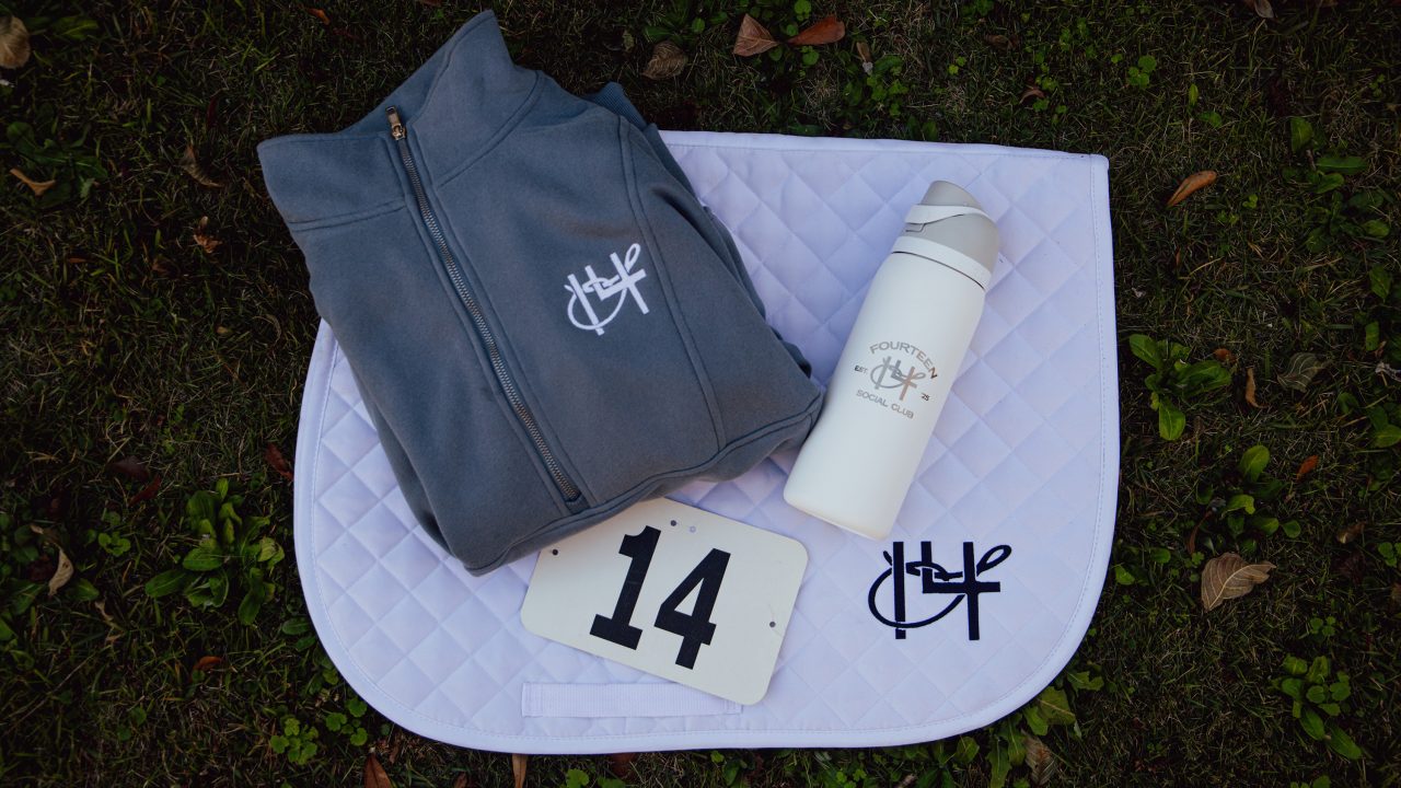 A grey jacket, a white water bottle, a white saddle pad, and a number 14 card are arranged on grass. All items display a matching equestrian-themed logo.