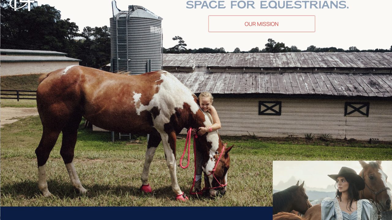 A young girl stands beside a brown and white horse near a barn, holding its reins as the horse grazes on grass.